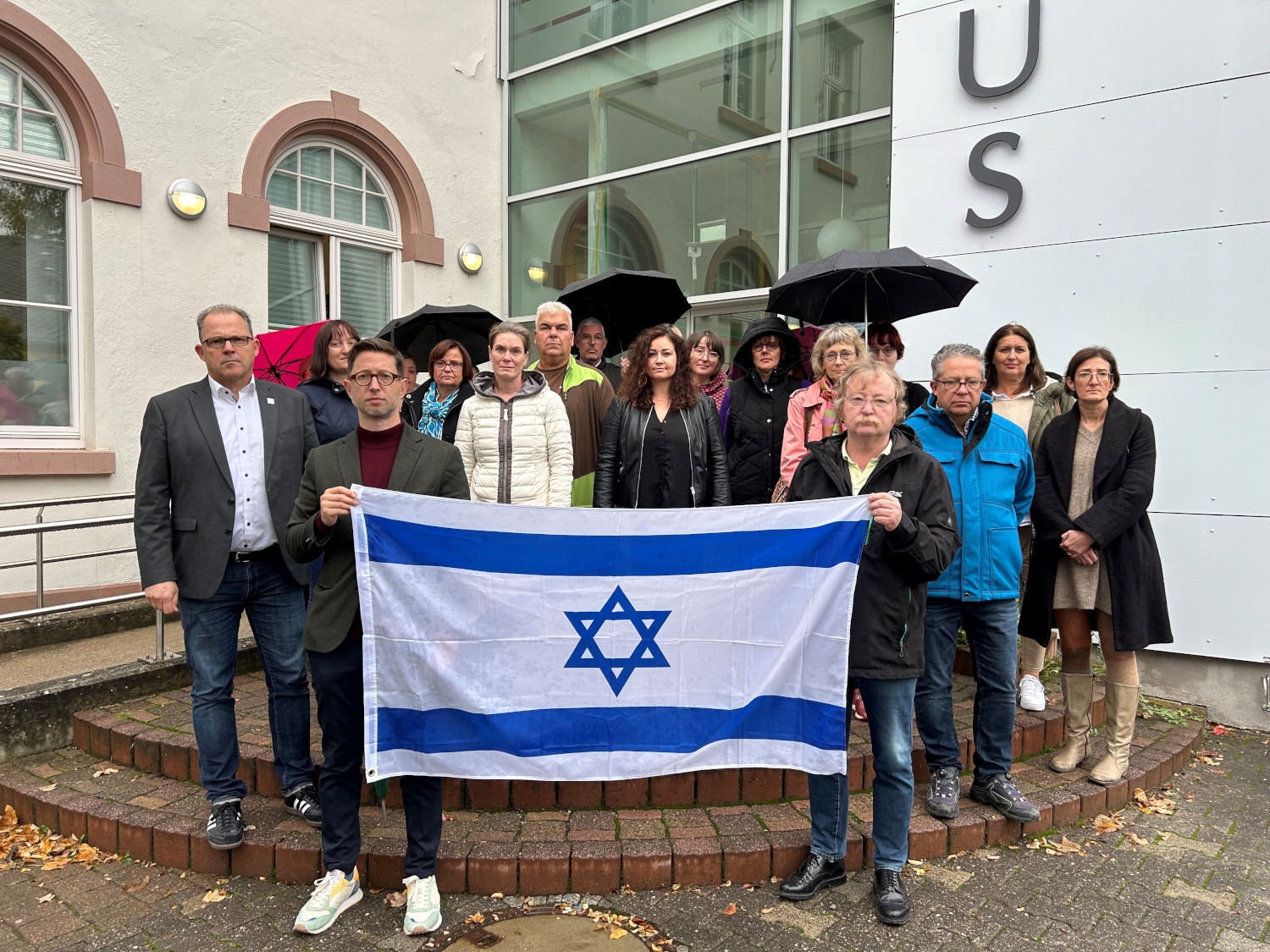 18 Personen stehen vor dem Rathaus Eltville und halten eine Flagge Israels vor sich.