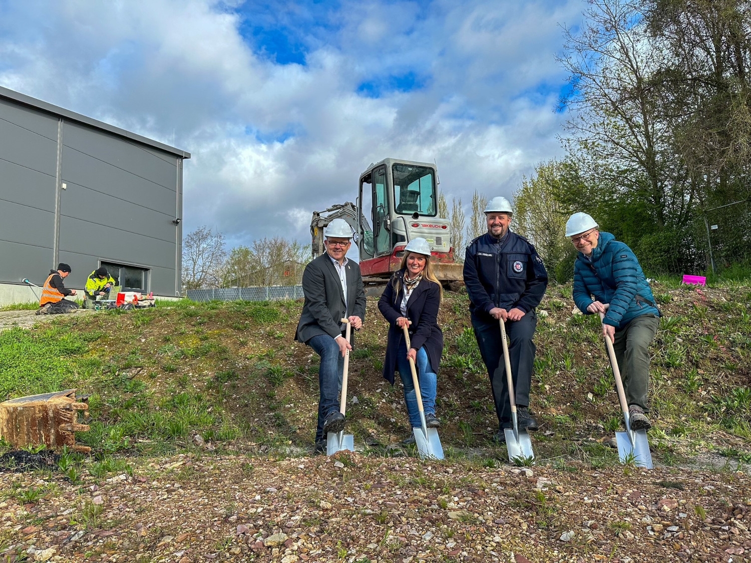 Die Beteiligten mit Bauhelmen und Spaten in der Hand, im Hintergrund arbeiten zwei Archäologen