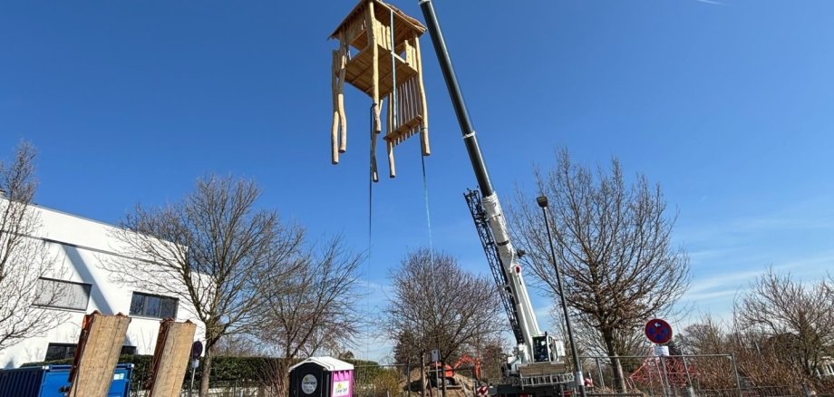 Ein Kran hebt einen Kletterturm aus Holz in die Luft.