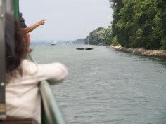 Blick auf den Rhein vom Schiff aus, eine Frau lehnt nach vorne gerichtet am Schiffsgeländer und jemand zeigt in die Ferne