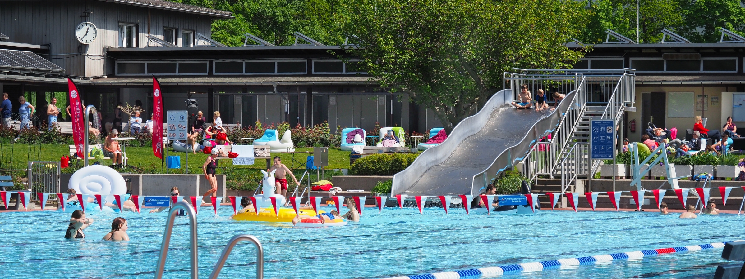 Immer was los im Eltviller Rosenbad. Schwimmerbecken mit rot-weißen-Fähnchen dekoriert. Im Hintergrund sieht man die Freibad-Rutsche und Personen, die im Wasser sind oder auf der Liegewiese entspannen.