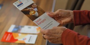Das Netzwerkbüro und MGH bieten ein vielfältiges Angebot an. Hände halten einen Flyer vom Sozialkomass, im HIntergrund liegen weitere Angebotsflyer.