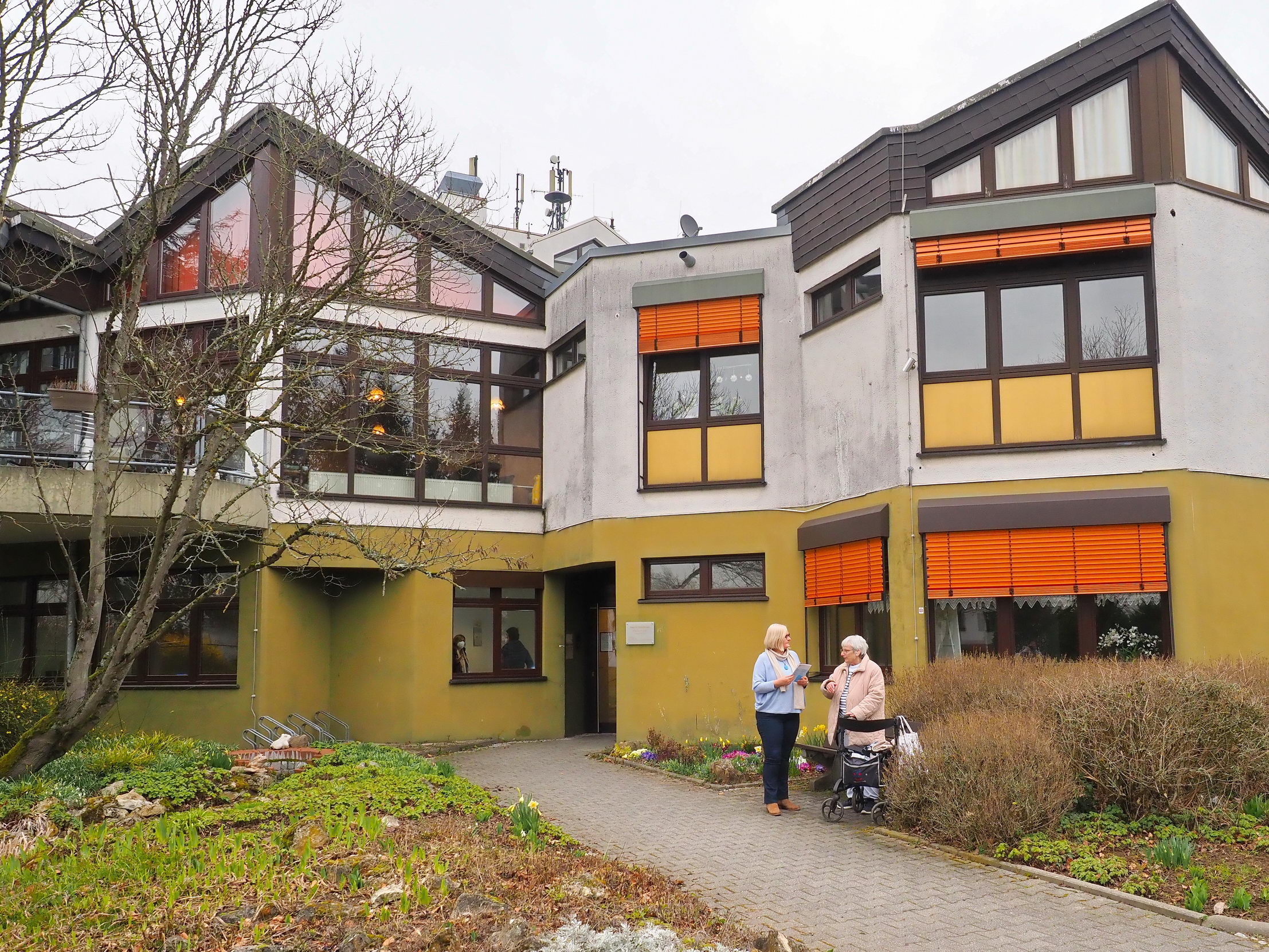 Ein Seniorenheim vom außen - davor stehen die Gemeindepflegerin und eine Seniorin.