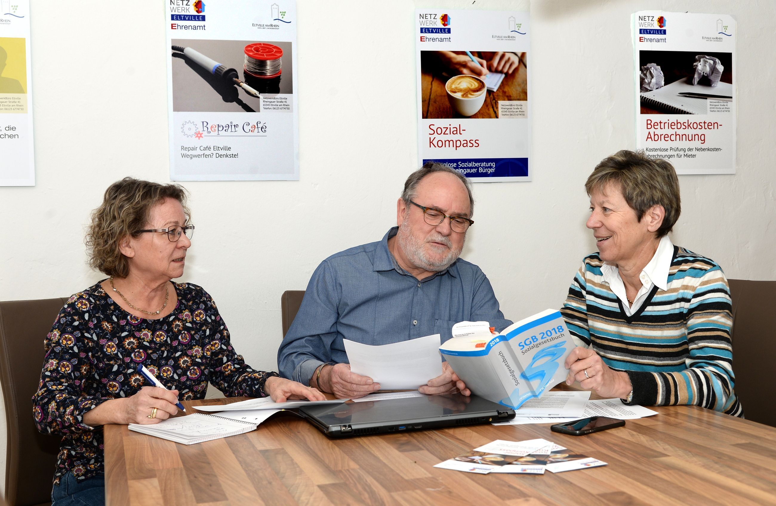 Das Team vom Rheingauer Sozialkompass kümmert sich um Ihre Anliegen. Ein Mann und zwei Frauen sitzen an einem Tisch und betrachten verschiedene Unterlagen. Eine Frau hält das Sozialgesetzbuch in der Hand.