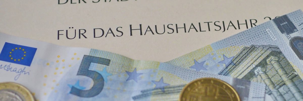 Ein fünf Euro Schein und mehrere Geldmünzen liegen auf einem Papier mit der Aufschrift "Haushaltsplan".