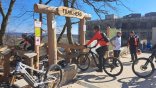 Man sieht den Eingang zur Mountainbike-Strecke, der aus großen Holzstämmen zusammen gebaut ist. Oben hängt ein Holzschild mit "Trailhead". Es sind vier Mountainbikes und zwei Fahrer zu erkennen im Hintergrund unterhalten sich zwei Männer. Ganz hinten ist die Fassade des WALD-WEIT Hotel zu erkennen.