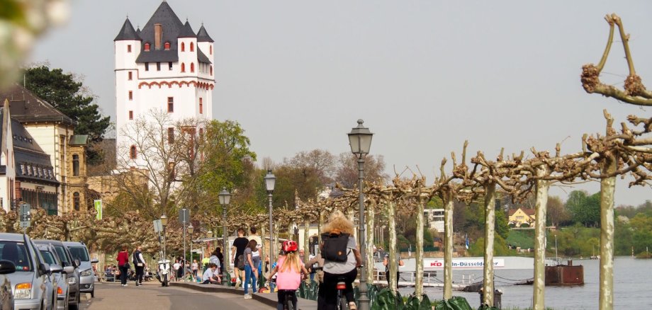 Eine Frau und ein Kind auf Fahrrädern fahren am Rheinufer in Richtung Burgturm.