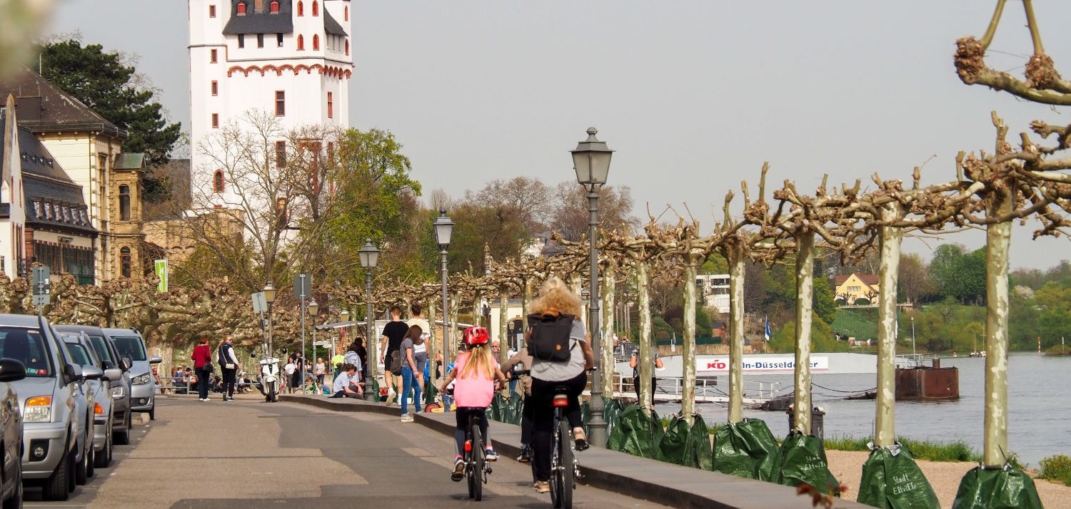 Eine Frau und ein Kind auf Fahrrädern fahren am Rheinufer in Richtung Burgturm.