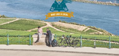 Eine Fahrradfahrerin und ein Farradfahrer stehen vor der Kulisse von Rhein und Weinbergen