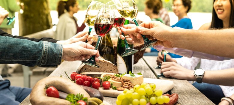 Vier Personen stoßen mit Weingläsern an, auf einem Tisch davor liegen drei Brotzeitteller.