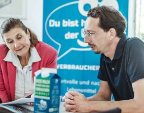 Eine Frau und ein Mann tauschen sitzen an einem Tisch und unterhalten mit verschiedenen Unterlagen vor sich, über die sie sich austauschen.