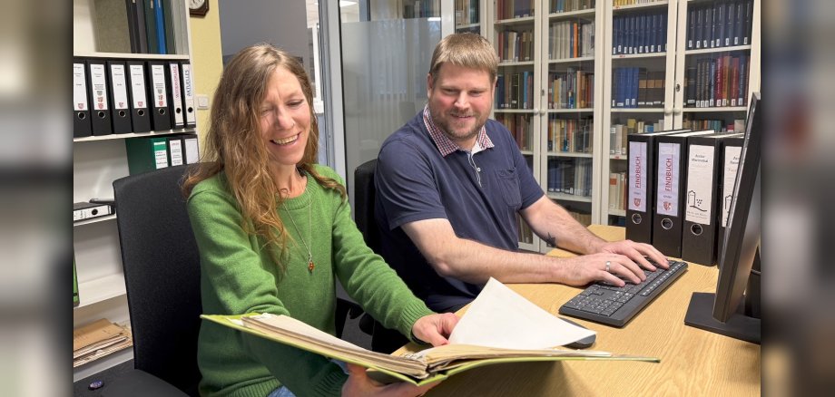 Mirjam Ehlers mit einem Findbuch in der Hand und Björn David beim Tippen auf der Tastatur