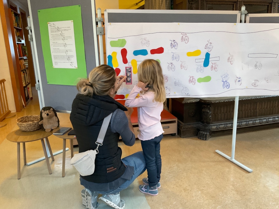 Mutter und Tochter von hinten zu sehen, wie sie ein Foto auf die gemeinsame Collage in der Kita kleben