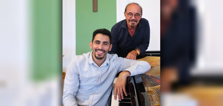 Aeham Ahmad sitzt am Klavier, Andreas Lukas steht hinter ihm, ebenfalls ans Klavier lehnend