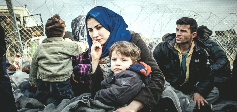 Frau mit Kopftuch schaut in die Kamera, sie hat ein Kleinkind im Arm; Mann und weiteres Kind im Hintergrund, hinter ihnen ist ein Zaun zu sehen